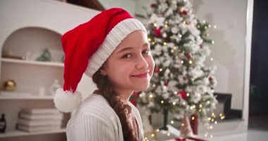 Noel Baba 'nın Noel ağacının yanındaki geleneksel oturma odasında oturmuş Noel Baba şapkalı gülümseyen mutlu çocuk portresi..