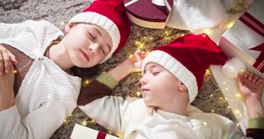 Kırmızı ve beyaz Noel şapkalı çocuklar tatlı rüyalar görür ve süslü geleneksel bir oturma odasında Noel hediyeleri kutusunun yanında yatarlar. Mutlu noeller. Mutlu yıllar 2025.