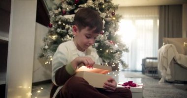 Noel hediyesi kutusunu açan yakışıklı ve şaşkın çocuk şöminenin yanındaki geleneksel oturma odasında oturuyor. Portre küçük çocuk, mutlu sürpriz çocuk