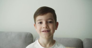Evdeki küçük okul çocuğunun portresi. Öğrenci, çocuk, yetim, çocuk, çocuk, yetim çocuk. Kanepede oturmuş kameraya gülümsüyor. 4K.