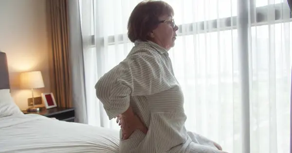 Kafkasyalı yaşlı bir kadın rahat bir yatak odasında yatağında sırt ağrısı çekiyor. Rahatlamak için sırtını iki eliyle tutuyor.