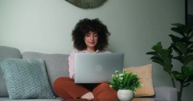 Kıvırcık saçlı güzel bir kadın rahat bir oturma odasında rahat bir koltukta oturuyor, dizüstü bilgisayarla çalışıyor ve rahat ve hoşnut görünüyor. Neşeli ve sakin görünüyor. Işıl ışıl bir gülümsemeyle. 4K