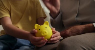 Çocuk, yakın planda kumbarada para biriktiriyor. Büyükannesi kumbaraya bozuk para atarken çocuk mavi kumbarasını elinde tutuyor. Çocuklar maddi olarak güvende. Finansal tasarruflar.