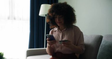 Evdeki koltukta oturan, elinde kredi kartı ve akıllı telefon tutan gülümseyen genç bayan müşteri. İnternetten alışveriş yapan mutlu bayan alışverişçi kolay taşınabilir ödemeler yapıyor. E-bankacılık uygulama servisi