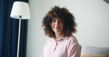 Kumral kıvırcık saçlı güzel bir kadın rahat rahat oturma odasındaki rahat koltukta oturuyor, kameraya mutlu bir şekilde gülümsüyor. Neşeli, sakin ve mutlu görünüyor. Sağlıklı, ışıl ışıl bir gülümsemeyle.