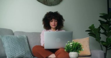 Kıvırcık saçlı güzel bir kadın rahat bir oturma odasında rahat bir koltukta oturuyor, dizüstü bilgisayarla çalışıyor ve rahat ve hoşnut görünüyor. Neşeli ve sakin görünüyor. Işıl ışıl bir gülümsemeyle. 4K