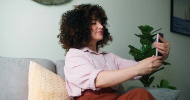 Kıvırcık saçlı güzel esmer kadın rahat oturma odasında rahat bir kanepeye oturur, selfie çeker ve kameraya mutlu bir şekilde gülümser. Kız selfie çekiyor mutlu görünüyor, neşeli, sakin, ışıl ışıl bir gülümsemeyle.