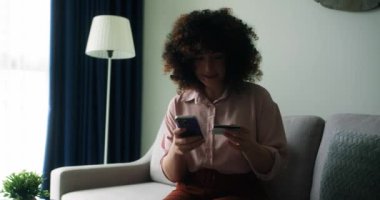 Evdeki koltukta oturan, elinde kredi kartı ve akıllı telefon tutan gülümseyen genç bayan müşteri. İnternetten alışveriş yapan mutlu bayan alışverişçi kolay taşınabilir ödemeler yapıyor. E-bankacılık uygulama servisi