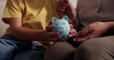 Çocuk, yakın planda kumbarada para biriktiriyor. Büyükannesi kumbaraya bozuk para atarken çocuk mavi kumbarasını elinde tutuyor. Çocuklar maddi olarak güvende. Finansal tasarruflar.