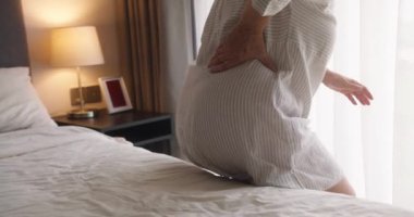 Kafkasyalı yaşlı bir kadın rahat bir yatak odasında yatağında sırt ağrısı çekiyor. Rahatlamak için sırtını iki eliyle tutuyor.