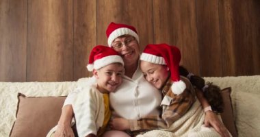Noel 'de torunlarıyla birlikte güzel dekore edilmiş bir Noel ağacının önünde, oturma odasında mutlu bir büyükanne. Büyükannem Noel Baba şapkası takarak torununa sarılıyor.