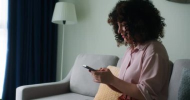 Oturma odasındaki kadın telefonuna zum yapıyor. Evde cep telefonuyla sosyal medya sohbeti yapan bir kız. Kikirdeyen kız yüzü. Kanepede cep telefonu ekranı yazarak iletişim kuruyor.