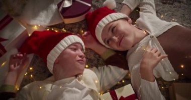 Mutlu noeller. Mutlu yıllar 2025. Kırmızı Noel Baba şapkalı çocuklar yeni yıl atmosferinde konuşur, çocuklar süslü geleneksel bir oturma odasında Noel hediyeleri kutusunun yanında yatar..