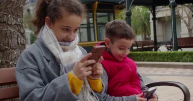 Parktaki bankta oturan çocuklar, telefonlarıyla oynuyorlar. Bir kız ve bir oğlan parkta bir bankta oturuyorlar, aletlerine, sosyal medyasına, modern çocuklara ve teknolojiye dalmışlar.