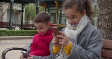 Parktaki bankta oturan çocuklar, telefonlarıyla oynuyorlar. Bir kız ve bir oğlan parkta bir bankta oturuyorlar, aletlerine, sosyal medyasına, modern çocuklara ve teknolojiye dalmışlar.