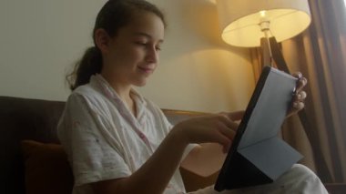 Çocuk tablette oyun oynuyor. Çocuk pijaması giymiş, akşamları evdeki oturma odasındaki kanepede oturan kız, evdeki odasındaki tableti kullanıyor.