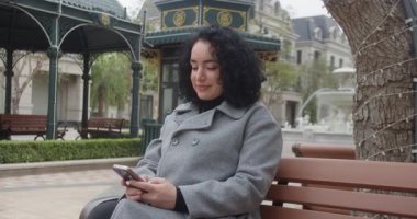 Rahat ceketli genç bir kadın gülüyor ve akıllı telefonuyla yazı yazıyor. Parktaki bir bankta sonbahar güzelliğiyle çevrili bir fıskiyenin yanında oturmuş mesajlar okuyor..