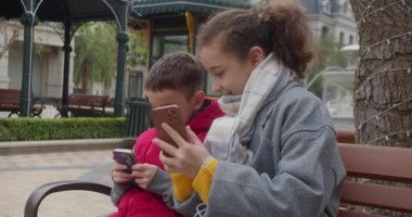 Parktaki bankta oturan çocuklar, telefonlarıyla oynuyorlar. Bir kız ve bir oğlan parkta bir bankta oturuyorlar, aletlerine, sosyal medyasına, modern çocuklara ve teknolojiye dalmışlar.