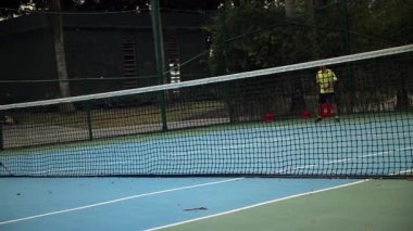 Tenis mavisi giymiş Asyalı yarışçılar sahada vakit geçiriyorlar, güneşli bir günde tenis oynuyorlar, tenis raketi tutuyorlar, yavaş hareketlerle tenis topuna vuruyorlar..