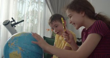 Mutlu çocuk ve kız büyüteçten dünyaya bakıyorlar. Okul çocukları sabah ışığında gülümserler evdeki bir masada oturup büyüteçle küreye bakarlar. 4K.