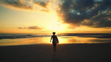 Sıcak okyanus sularında turuncu bir yaz günbatımında yürüyen, deniz kıyısında bir köpekle koşan seksi, ince kadın silueti. Pelerinli seksi, baştan çıkarıcı siluet kız gün batımında köpekle deniz kenarında yürüyor..