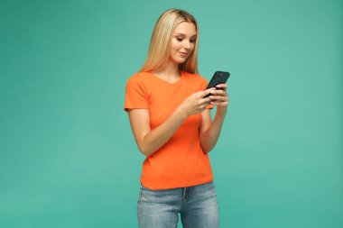 Turuncu tişörtlü, kot pantolonlu, siyah telefonlu güzel sarışın kız. Mavi arka plan. Stüdyo.