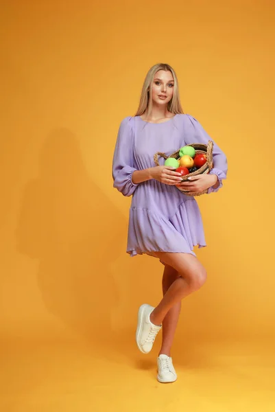Beautiful blonde girl in a lilac dress with a basket of fruits on a yellow background. Studio. Healthy.