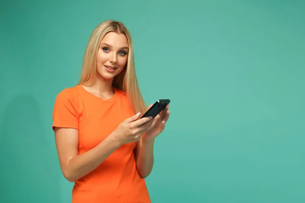 Beautiful blonde girl in an orange t-shirt and jeans with a black phone. Blue background. Studio.