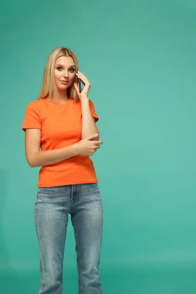 Beautiful blonde girl in an orange t-shirt and jeans with a black phone. Blue background. Studio.