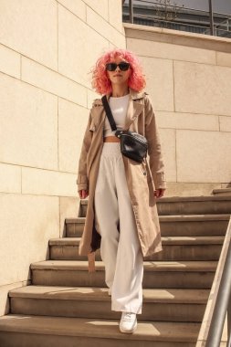 Pembe saçlı şık bir kızın portresi. Bej gözlükler. Bahar ve yaz trendi 2023.Bej Çukurlu pelerin beyaz bluz ve pantolonlu. Siyah küçük kemer çantası.