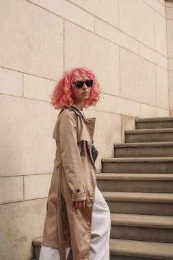 Pembe saçlı şık bir kızın portresi. Bej gözlükler. Bahar ve yaz trendi 2023.Bej Çukurlu pelerin beyaz bluz ve pantolonlu. Siyah küçük kemer çantası.