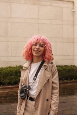 Pembe saçlı şık bir kızın portresi. Bej gözlükler. İlkbahar ve yaz trendi 2023. Beyaz tişört ve bej pantolonlu bej siper pelerini. Siyah küçük kemer çantası.