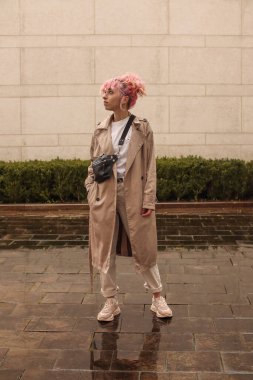 Pembe saçlı şık bir kızın portresi. Bej gözlükler. İlkbahar ve yaz trendi 2023. Beyaz tişört ve bej pantolonlu bej siper pelerini. Siyah küçük kemer çantası.