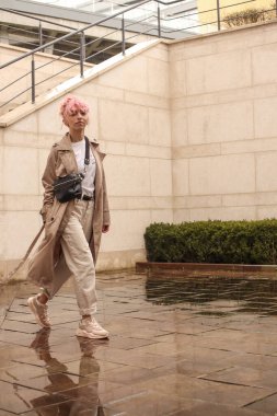 Pembe saçlı şık bir kızın portresi. Bej gözlükler. İlkbahar ve yaz trendi 2023. Beyaz tişört ve bej pantolonlu bej siper pelerini. Siyah küçük kemer çantası.