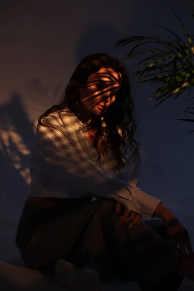 一个穿着白衬衫和深色裤子的时髦黑发女孩坐在白色的背景上 一束光线照在脸上 流行的波荷风格的枝条 棕榈叶的阴影 — 图库照片