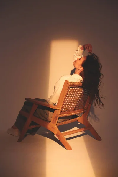 一个穿着白衬衫和深色裤子的时髦黑发女孩坐在白色的背景上 一束光线照在脸上 流行的波荷风格的枝条 棕榈叶的阴影 — 图库照片