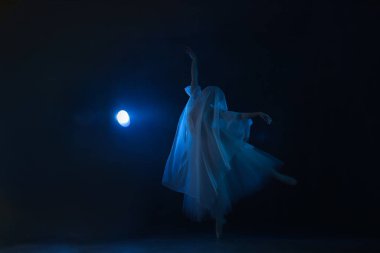 Profesyonel balerin dans ediyor. Beyaz elbiseli ve sivri uçlu ayakkabılı balerin. Koyu arkaplan, mavi ışık. Peçe. Sahne. Performans