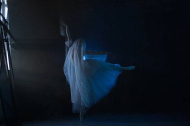 Profesyonel balerin dans ediyor. Beyaz elbiseli ve sivri uçlu ayakkabılı balerin. Koyu arkaplan, mavi ışık. Peçe. Sahne. Performans