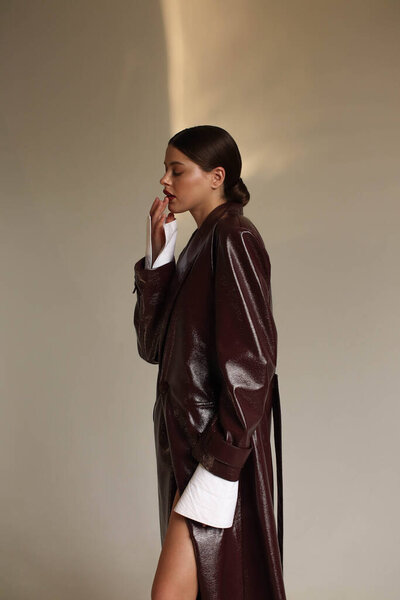 Portrait shooting of a stylish European girl in the studio. Dark hair and red lips. Model in a bodysuit and a long burgundy leather raincoat and sandals. Beige background. Fashion & Catalogs. Clothing trends for spring and autumn,trench