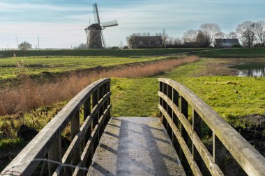 Ufukta geleneksel yel değirmeni olan yemyeşil bir çayırda büyüleyici bir köprünün olduğu sakin bir kış sahnesi. Kırsal sadeliğin ve zamansız güzelliğin bir görüntüsü.
