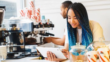 Önlüklü mutlu kadın internetten sipariş vermek için tablet kullanıyor, adam arka planda kahve yapıyor. Barista ve garson servisi konsepti