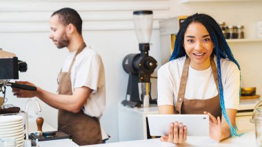 Kahve dükkanında çalışan mutlu bir bayan dijital tablet tutarken internetten sipariş alıyor. Erkek barista arka planda kahve yapıyor.