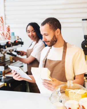 Barista internetten sipariş verirken dijital tablet kullanıyor ve elinde kahve çekirdekleri var. Kadın barista arka planda kahve yapıyor.