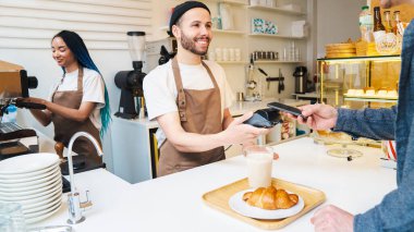 NFC teknolojisiyle ödeme yapan bir erkek müşteri kafede telefonla konuşuyor. Gülümseyen barista duruş terminalini tutuyor. Dijital ödeme kavramı