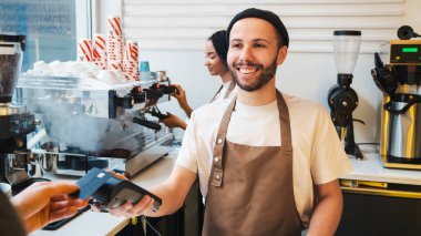 Kahve dükkanındaki müşteriden ödeme alırken elinde kredi kartı makinesi olan mutlu bir erkek barista. Arka planda kahve yapan kadın.