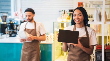 Gülümseyen garson elinde boş ekranla tablet, erkek barista elinde kahve çekirdekleri olan kağıt paketle ayakta duruyor. Boşluğu kopyala