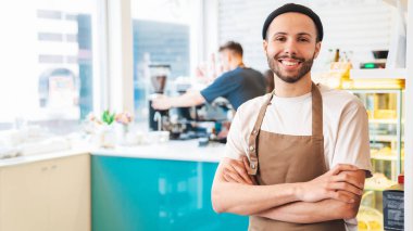Kollar bağlıyken kameraya bakarak gülümseyen şık erkek barista. Arka planda kahve yapan erkek arkadaş. Boşluğu kopyala