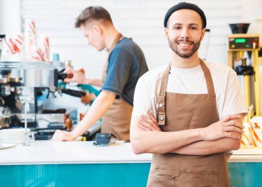 Kahve dükkanında çalışan mutlu, şık bir adam barın önünde duruyor, kollarını kavuşturuyor. Erkek meslektaşım arka planda kahve yapıyor.