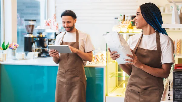Smiling waitress with package with coffee beans while male barista accepting online order while using digital tablet. Purchase concept