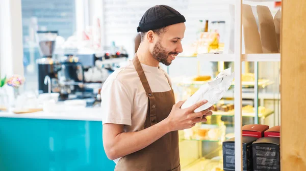 Smiling male barista reading the label on the packaging of coffee beans while standing near the rack. Business restaurant food and drink concept